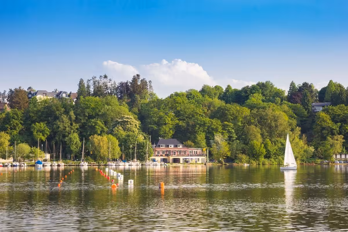 Segelboot auf ruhigem See vor bewaldeter Uferlandschaft.