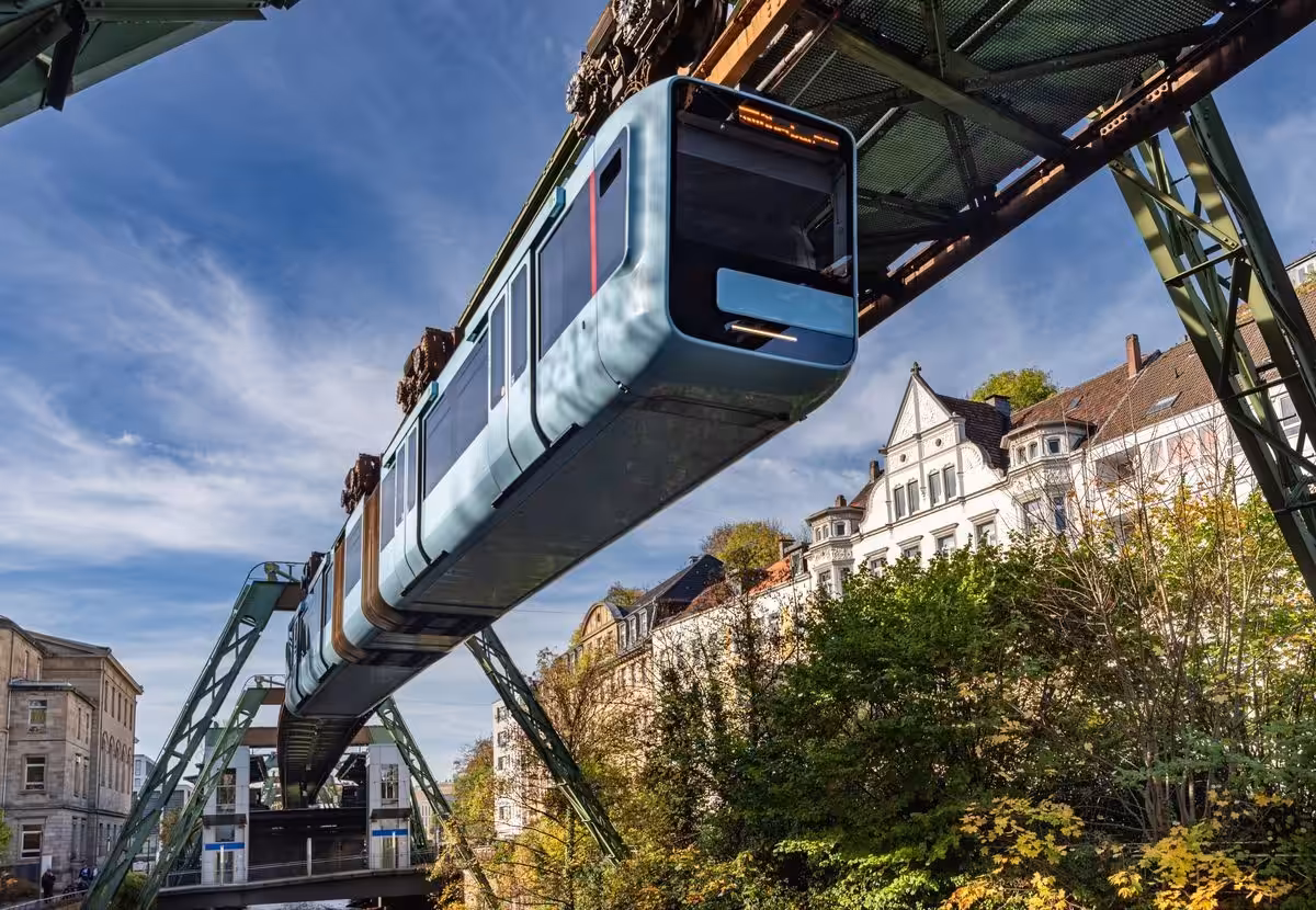 Wuppertaler Schwebebahn überquert Fluss vor Gebäuden.